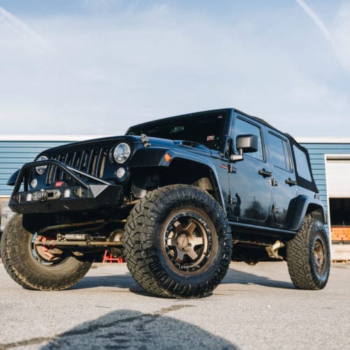 Black 4 dour soft top jeep with offroad style front bumper rotiform wheels and Nitto tires low front driver side grille view