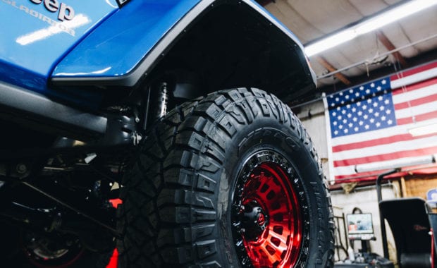 Blue Jeep Gladiator soft top with Red fuel wheels on lift in shop with American flag in background